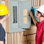 Two construction workers wearing hard hats examine an electrical panel in a building
