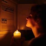 Young woman with candles in complete darkness examines fuse box or electrical distribution board at home