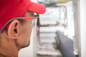 Caucasian Electrician Wearing Safety Glasses Looking Inside Electric Breaker Board For Potential Issues. Residential Electric Circuit.
